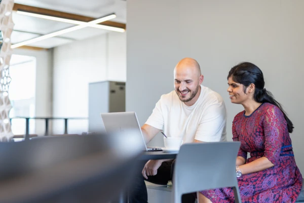 Dos compañeros sonríen mientras revisan contenidos en una tableta en una oficina moderna, como muestra de trabajo en equipo y colaboración.