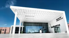 Lối vào hiện đại của Koelnmesse với những cánh cửa kính dưới mái che màu trắng, bầu trời xanh trong vắt và các biển chỉ dẫn “Eingang Süd” và “Entrance South”.