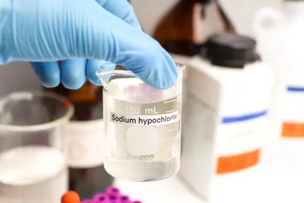 Hand holding a beaker labeled "Sodium hypochlorite" with a clear liquid, surrounded by laboratory bottles.