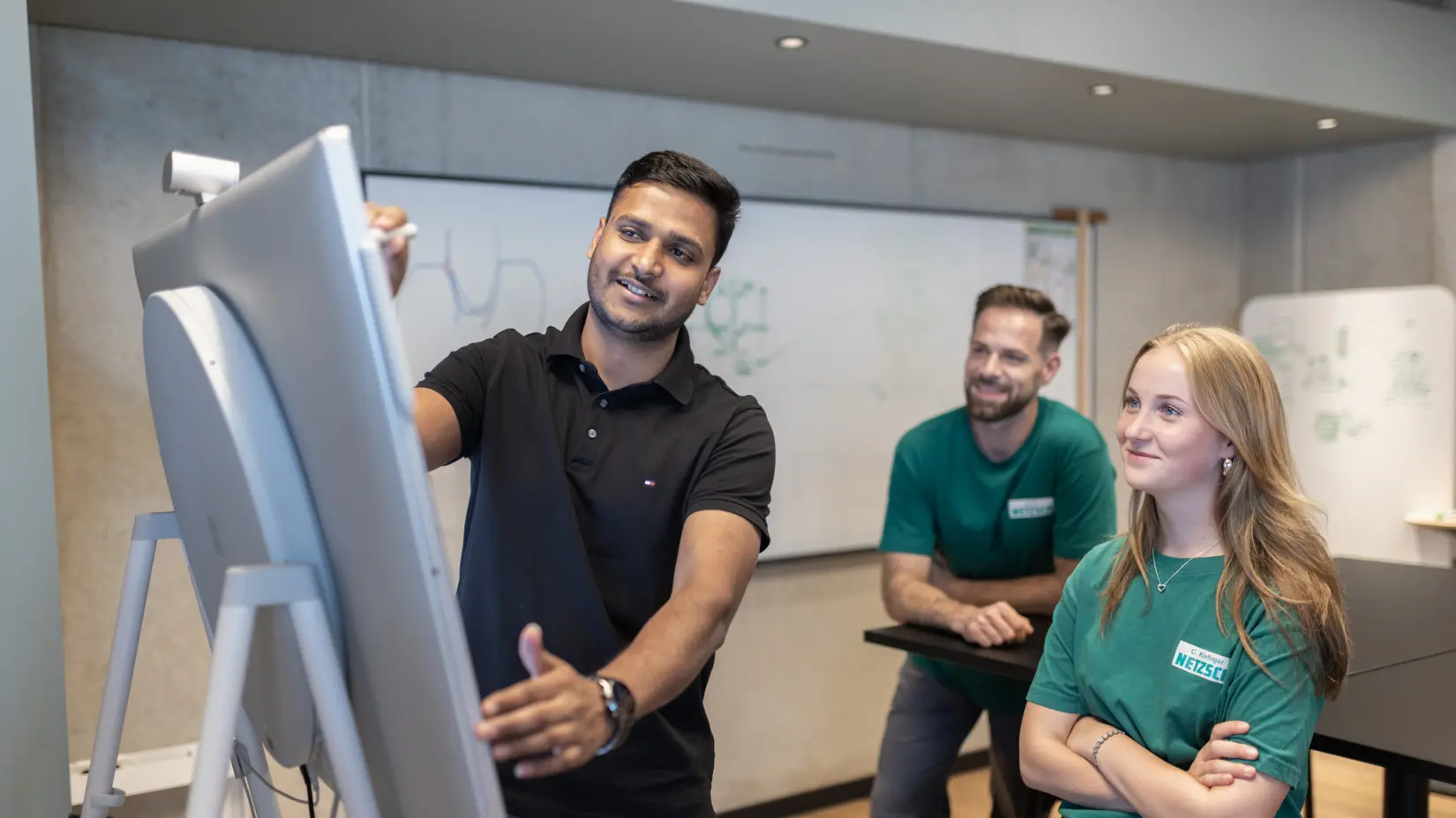 Equipe composta por profissionais de diversas áreas, trabalhando em conjunto em um escritório moderno, discutindo ideias com o auxílio de um quadro branco e de um monitor digital.