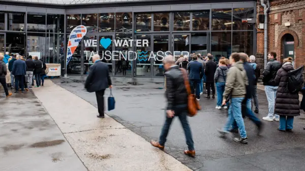 Tijdens een evenement in de watersector komen bezoekers een modern gebouw binnen met grote glazen ramen waarop „WE ♥ WATER TAUSENDWASSER ” staat.