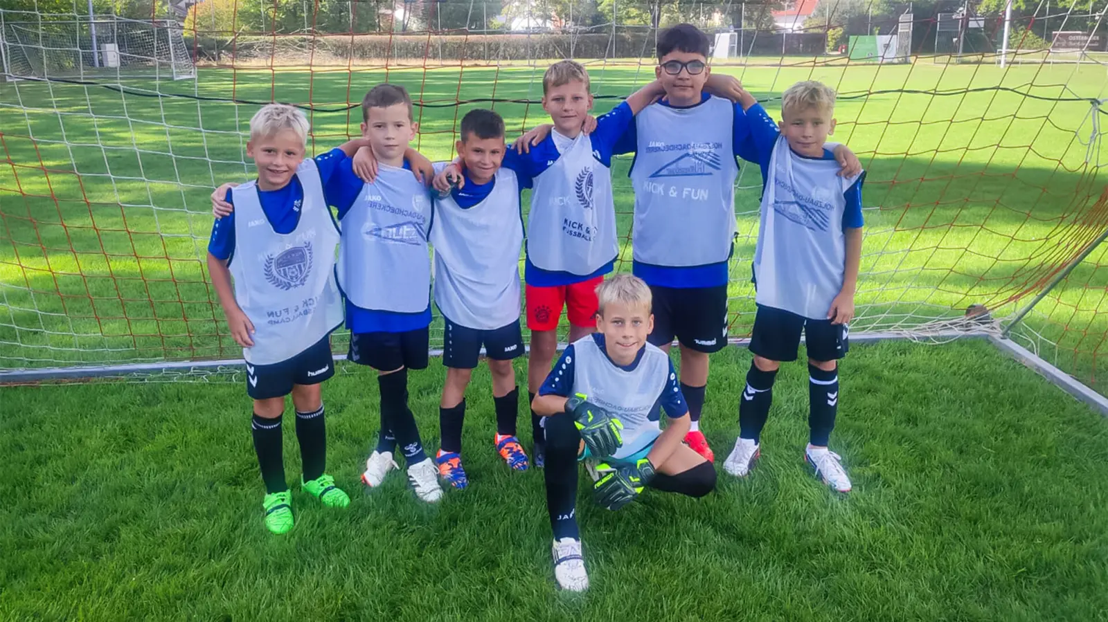 Sieben Kinder in blau-weißen Trikots posieren lachend vor einem Fußballtor auf grünem Rasen beim integrativen Kick & Fun Fußballcamp.