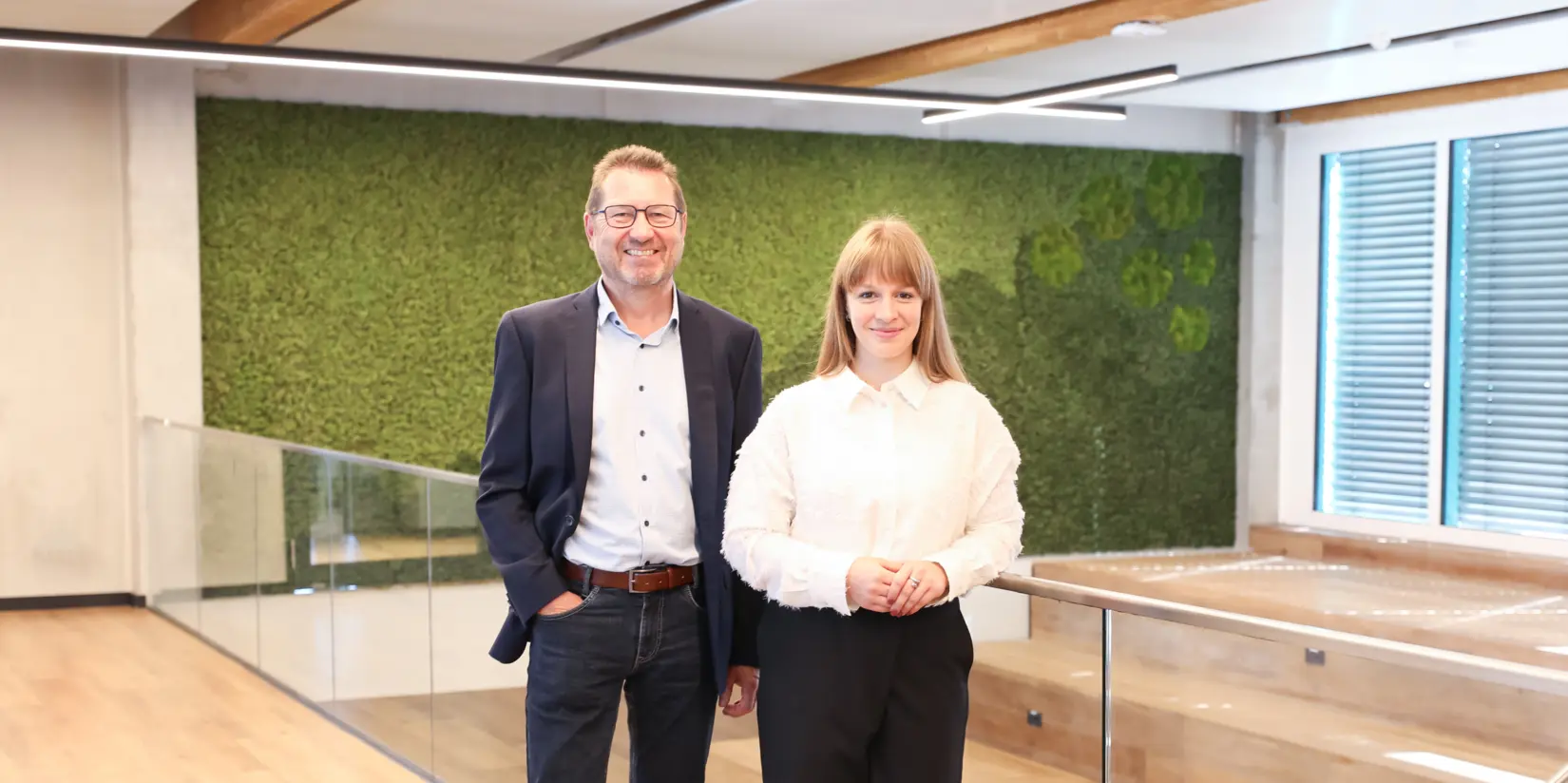 Laura Lex and Johann Vetter stand smiling in a modern, eco-friendly office space with wooden and green elements, emphasising sustainability at NETZSCH.