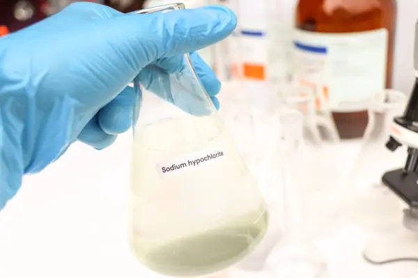 Una mano con un guanto che regge una fiaschetta con l’etichetta “ipoclorito di sodio”, circondata da vetreria da laboratorio e da un microscopio.