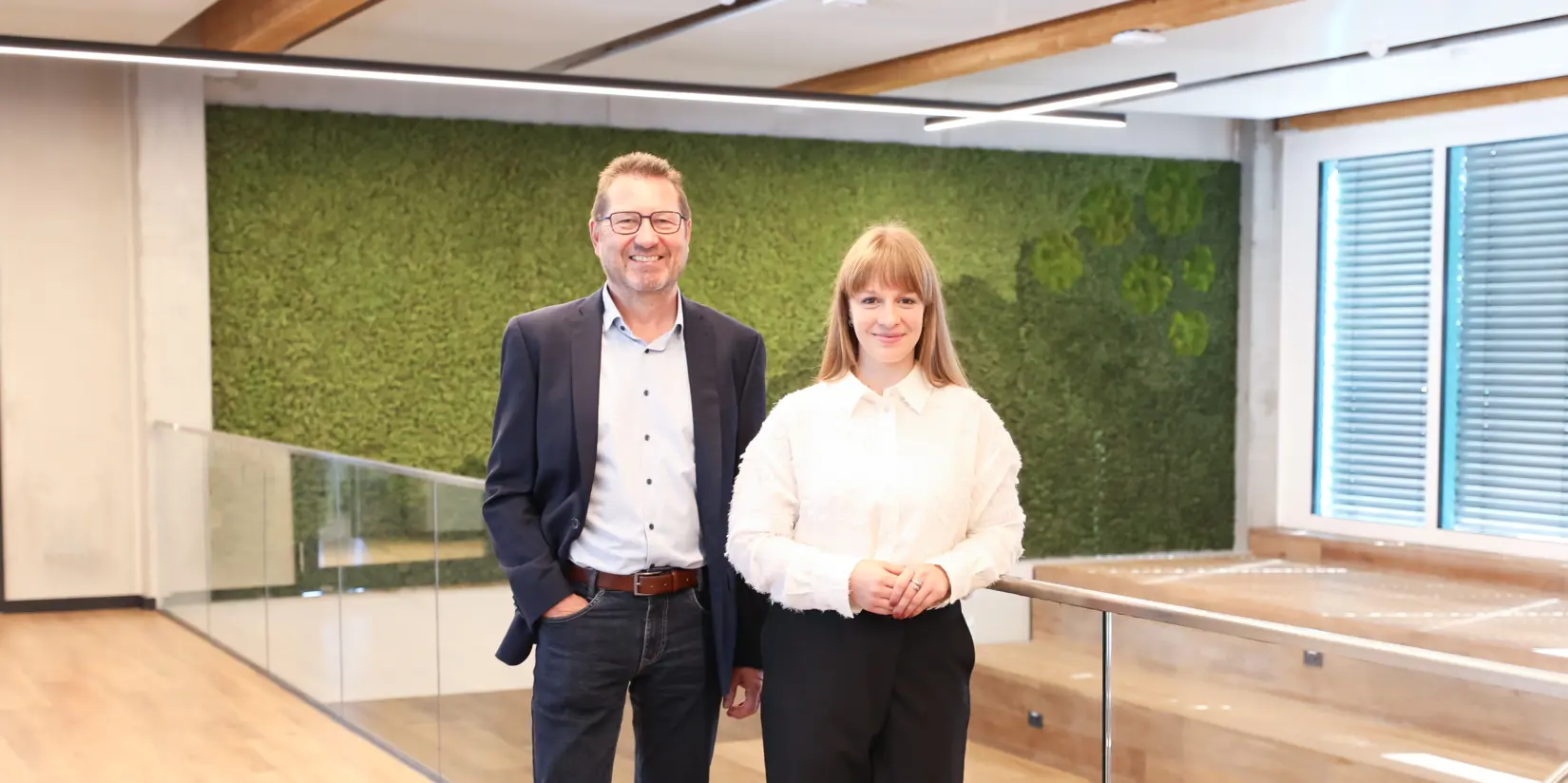 Laura Lex und Johann Vetter standen lächelnd in einem modernen, umweltfreundlichen Büroraum mit Holz- und Grünelementen, die die Nachhaltigkeit bei NETZSCH unterstreichen.