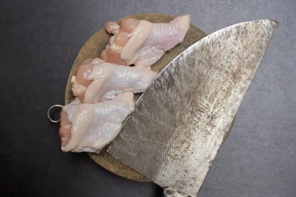 Alitas de pollo crudas colocadas junto a un cuchillo de carnicero grande y desgastado sobre una tabla de cortar redonda de madera, listas para su preparación.